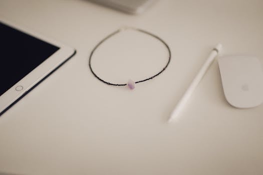 A stylish, minimalist workspace featuring a necklace, iPad, and Apple Pencil on a white surface.