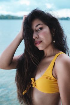 A woman with long hair poses confidently in a yellow bikini at the beach, exuding summer vibes.