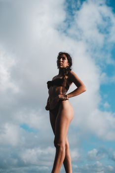 A woman confidently poses in a bikini against a backdrop of blue skies and white clouds.