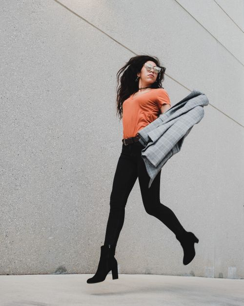 Free Woman in black leggings and orange top jumping gracefully against a concrete background. Stock Photo