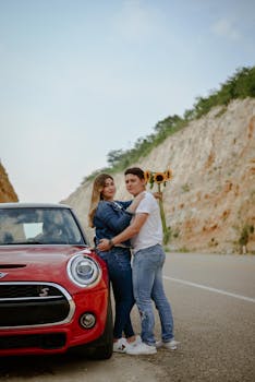A couple sharing a moment by their car on a scenic road, with sunflowers in hand, depicting love and togetherness.