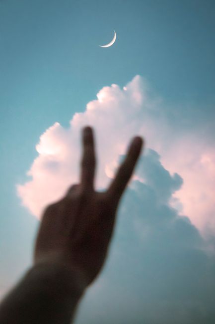Fotomural Silueta de una mano haciendo un signo de paz con luna creciente y nubes en un cielo azul.