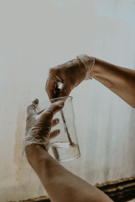 Hands in gloves stirring a liquid in a clear glass beaker with a neutral background.
