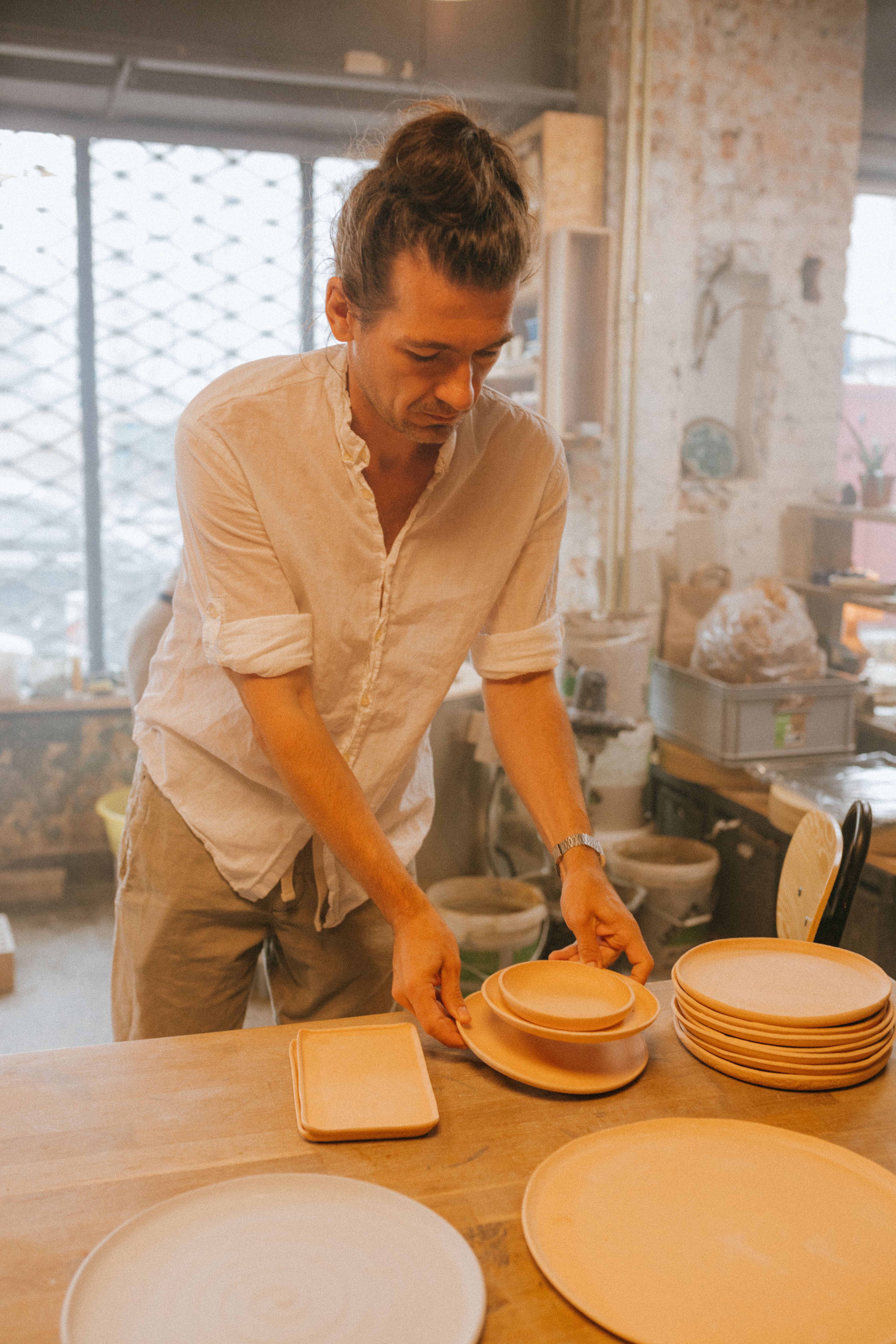 Man in white shirt arranging handmade ceramic plates in a sunlit studio.