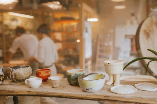 Free Ceramic bowls and cups in an artisan workshop with blurred figures in the background. Stock Photo