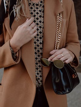 A fashionable woman in a coat holds sunglasses and a black bag outdoors.