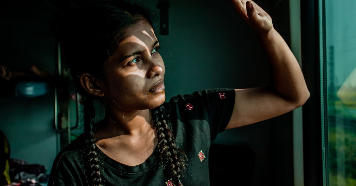 A woman with braided hair raises her hand towards the window, contemplating as light and shadow play on her face.