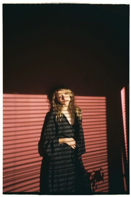 A woman in a robe stands indoors, highlighted by dramatic shadows from window blinds.