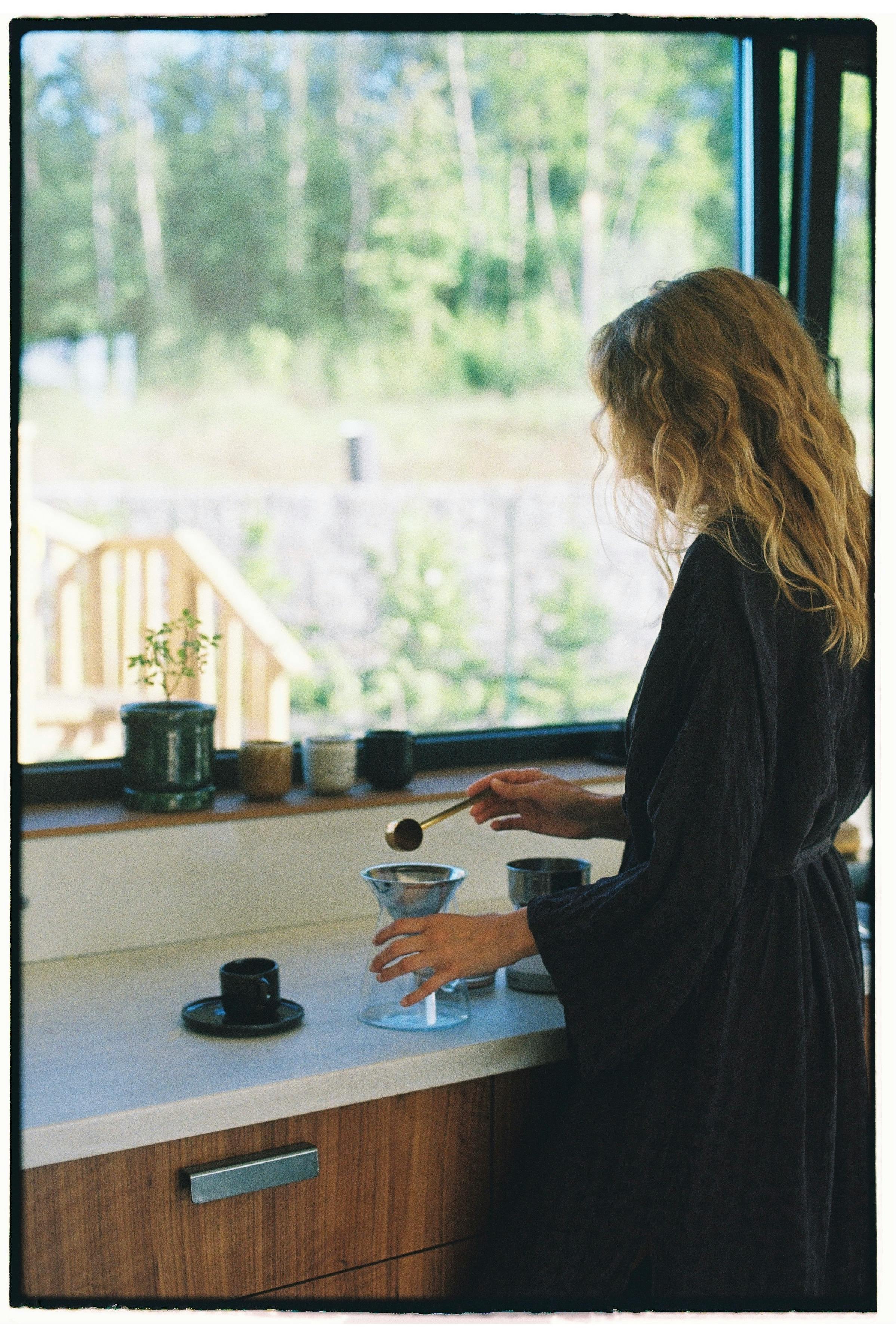 Blonde woman prepares pour over coffee in a sunlit kitchen.