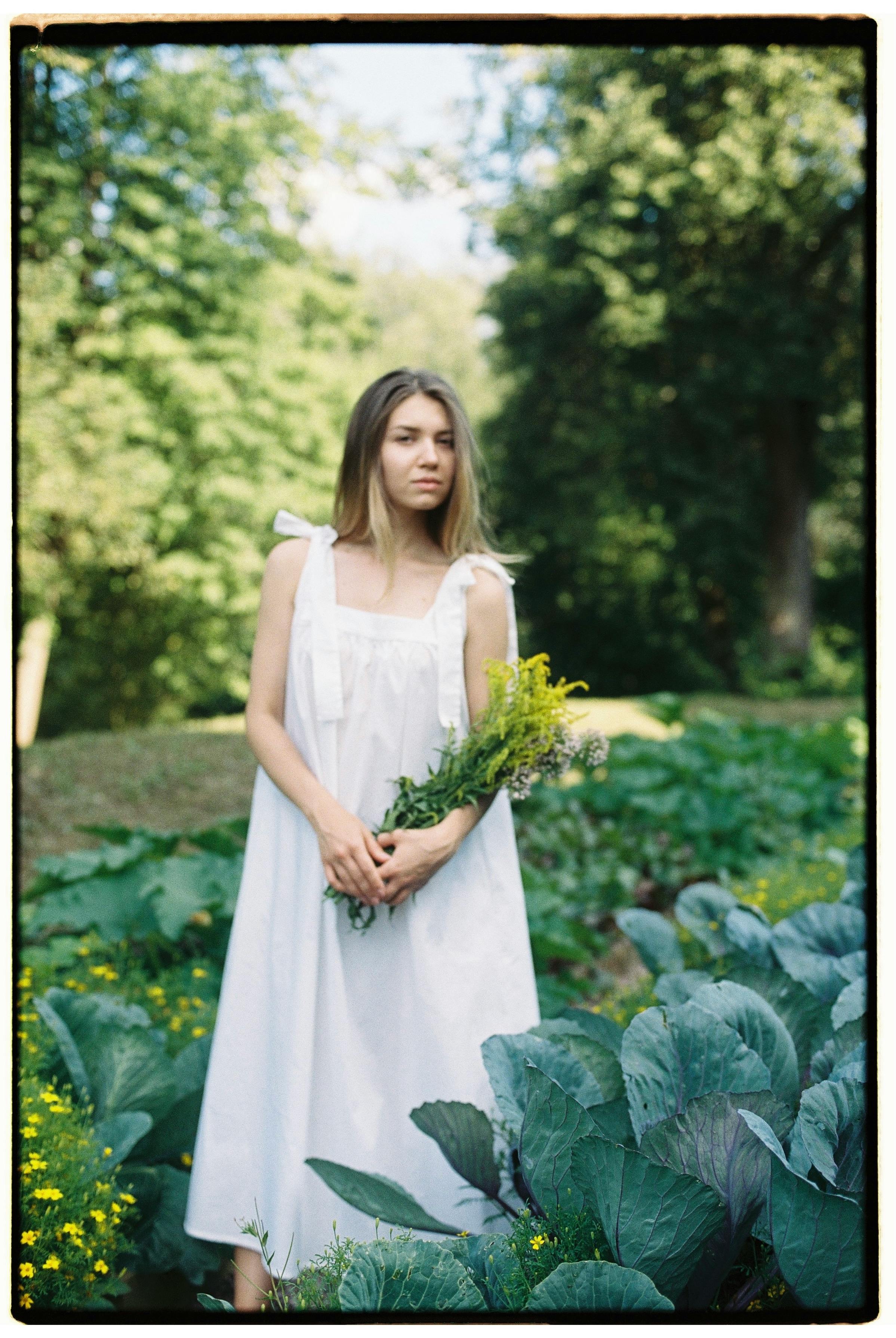 A young woman in a white dress stands in a lush garden holding flowers. Sunlight filters through the trees in this serene scene.