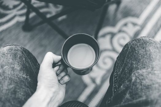 Black and white close-up of a hand holding a coffee cup over denim jeans.. black, white, close