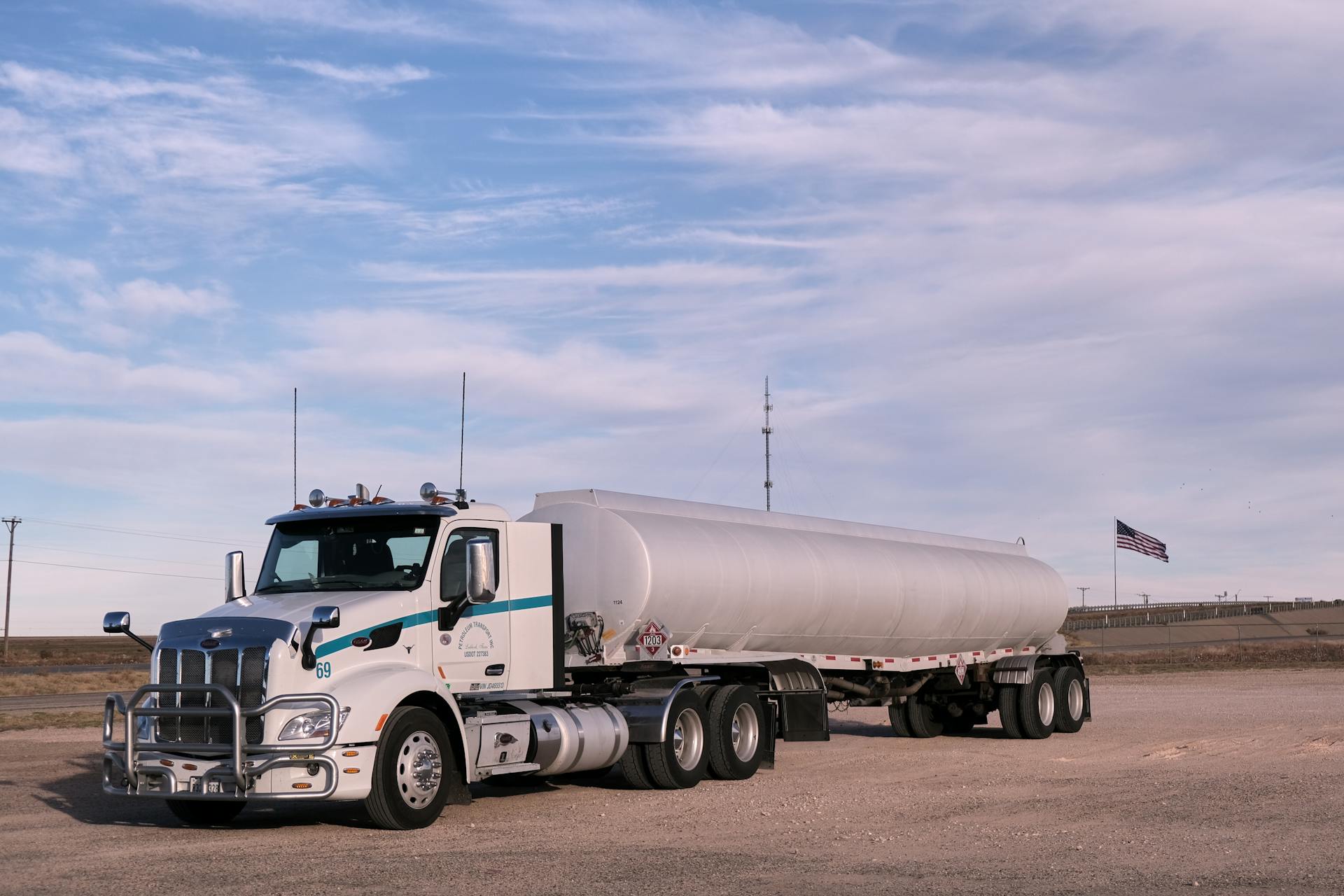 White fuel transport truck parked in a Texas landscape under clear skies, showcasing American flag in the distance.