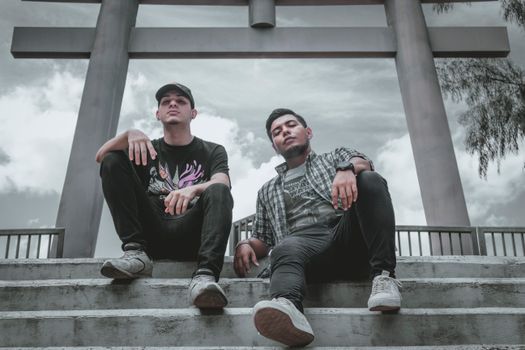 Two stylish young men casually posing on concrete steps outdoors under a cloudy sky.