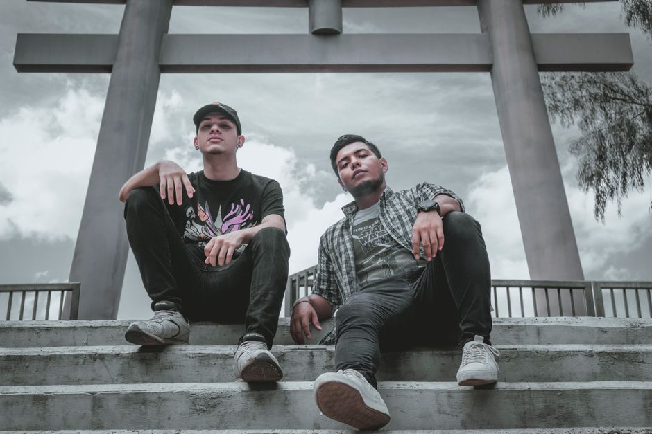 Two stylish young men casually posing on concrete steps outdoors under a cloudy sky.