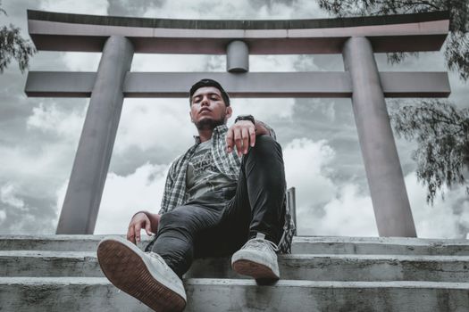 Casual young man in urban attire posing under a traditional Torii gate with cloudy skies.