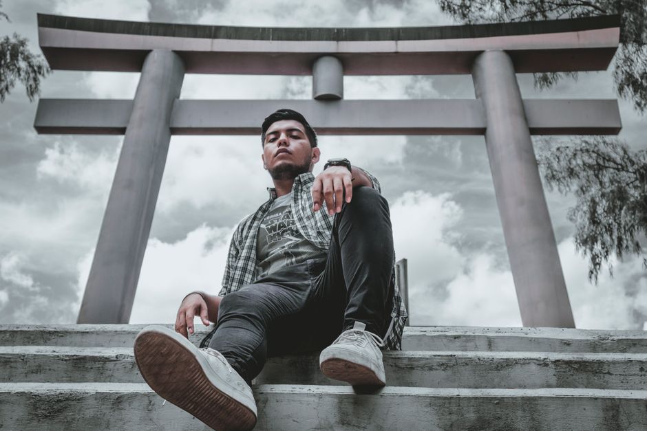 Casual young man in urban attire posing under a traditional Torii gate with cloudy skies.