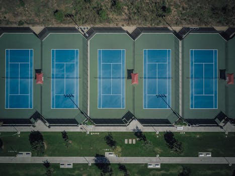A bird's-eye view of five outdoor tennis courts surrounded by greenery in a park setting.