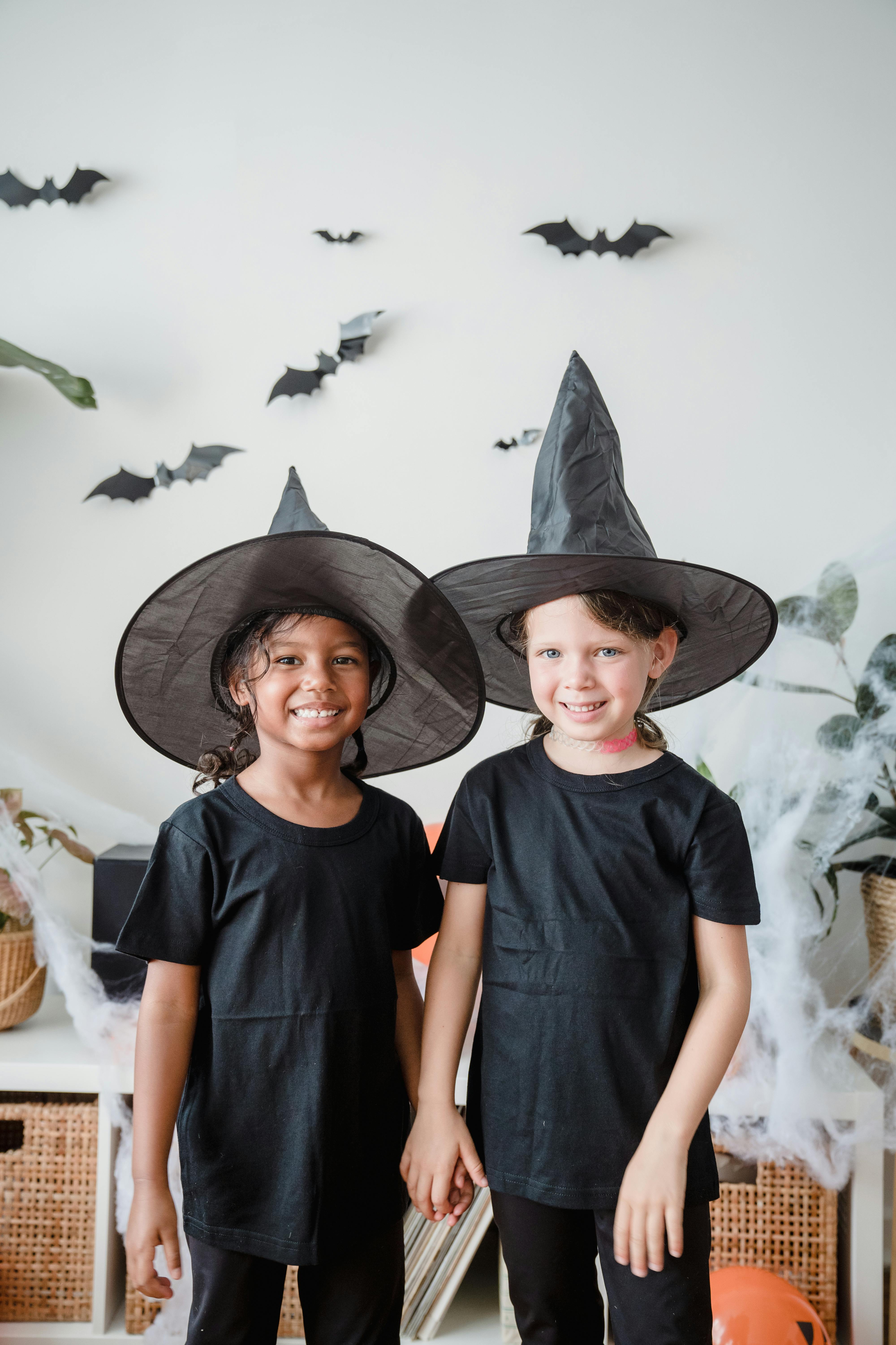 Two children dressed as witches, smiling and standing indoors decorated for Halloween.