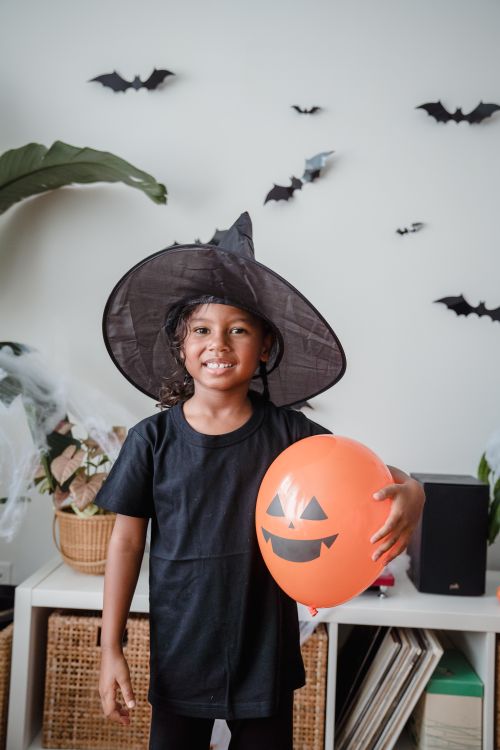 Free Happy child dressed as a witch, holding a pumpkin balloon, indoors on Halloween with paper bats decor. Stock Photo