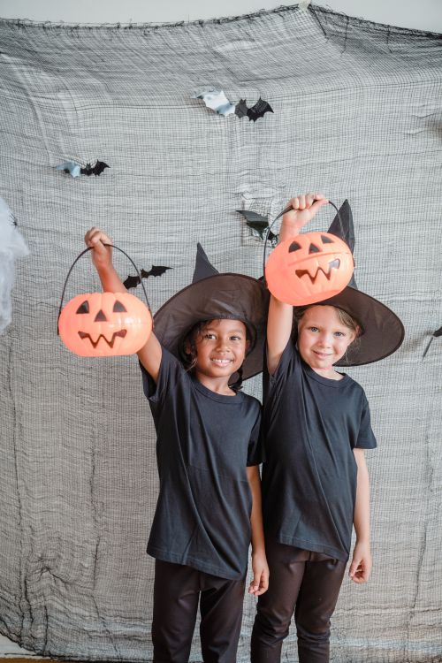 Free Two kids in witch hats celebrate Halloween indoors, holding jack-o'-lantern buckets with smiles. Stock Photo