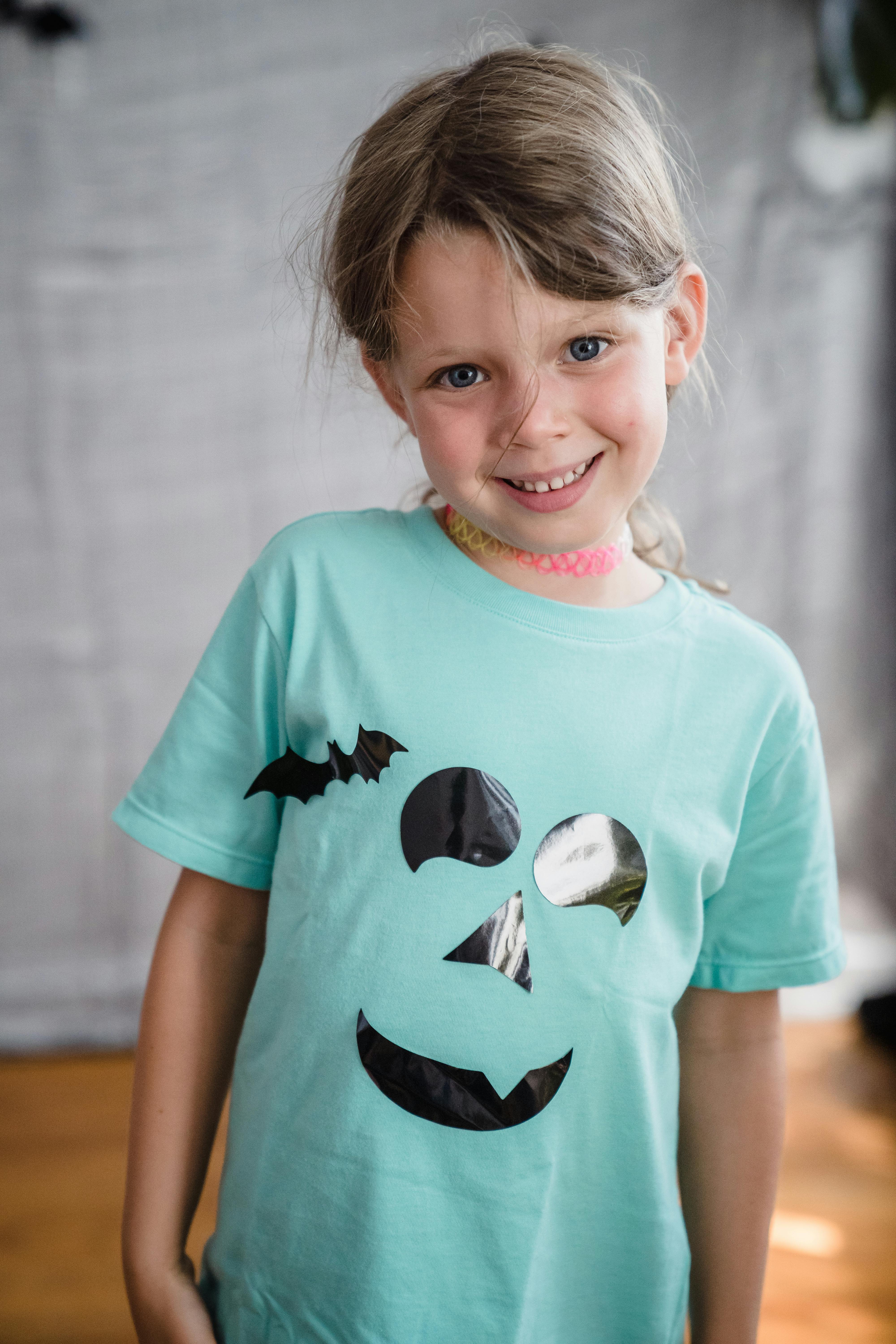 Young girl with brown hair smiling in a Halloween-themed t-shirt indoors.