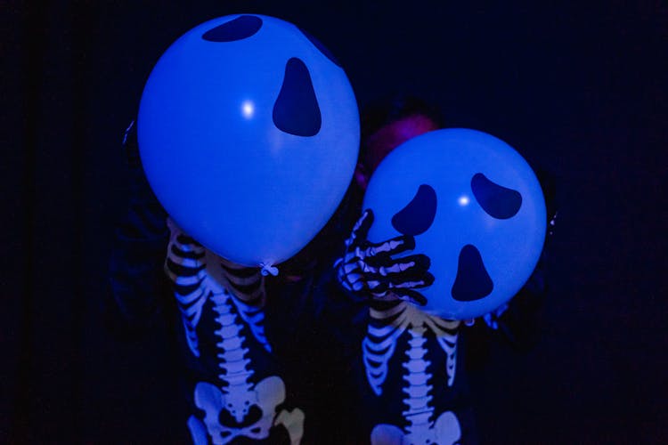 Two Kids Dressed As Skeletons Covering Faces With Balloons Under Blue Light