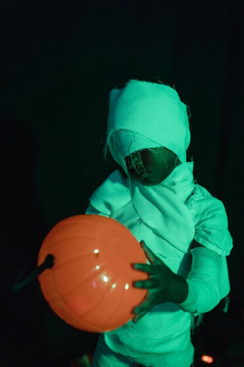 Free A child in a mummy costume holds a glowing pumpkin pail against a dark background. Stock Photo