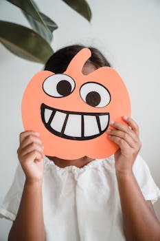 A child in a white shirt holding a cartoon pumpkin mask indoors.