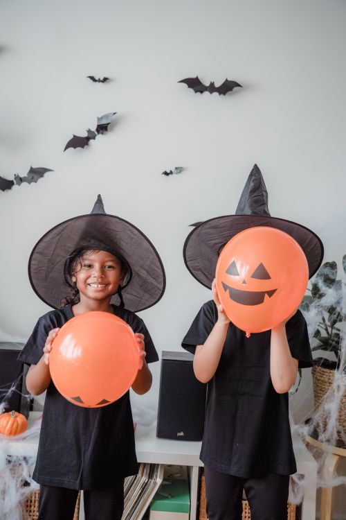 Free Kids dressed as witches holding orange balloons in a decorated Halloween setting. Stock Photo
