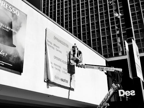 Free Grayscale Photo of Worker on an Aerial Lift Fixing a Billboard Stock Photo