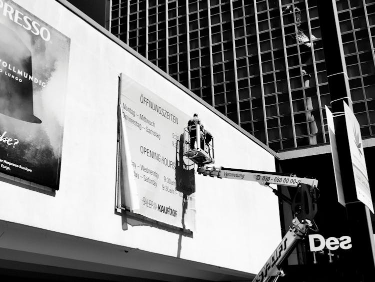 Grayscale Photo Of Worker On An Aerial Lift Fixing A Billboard