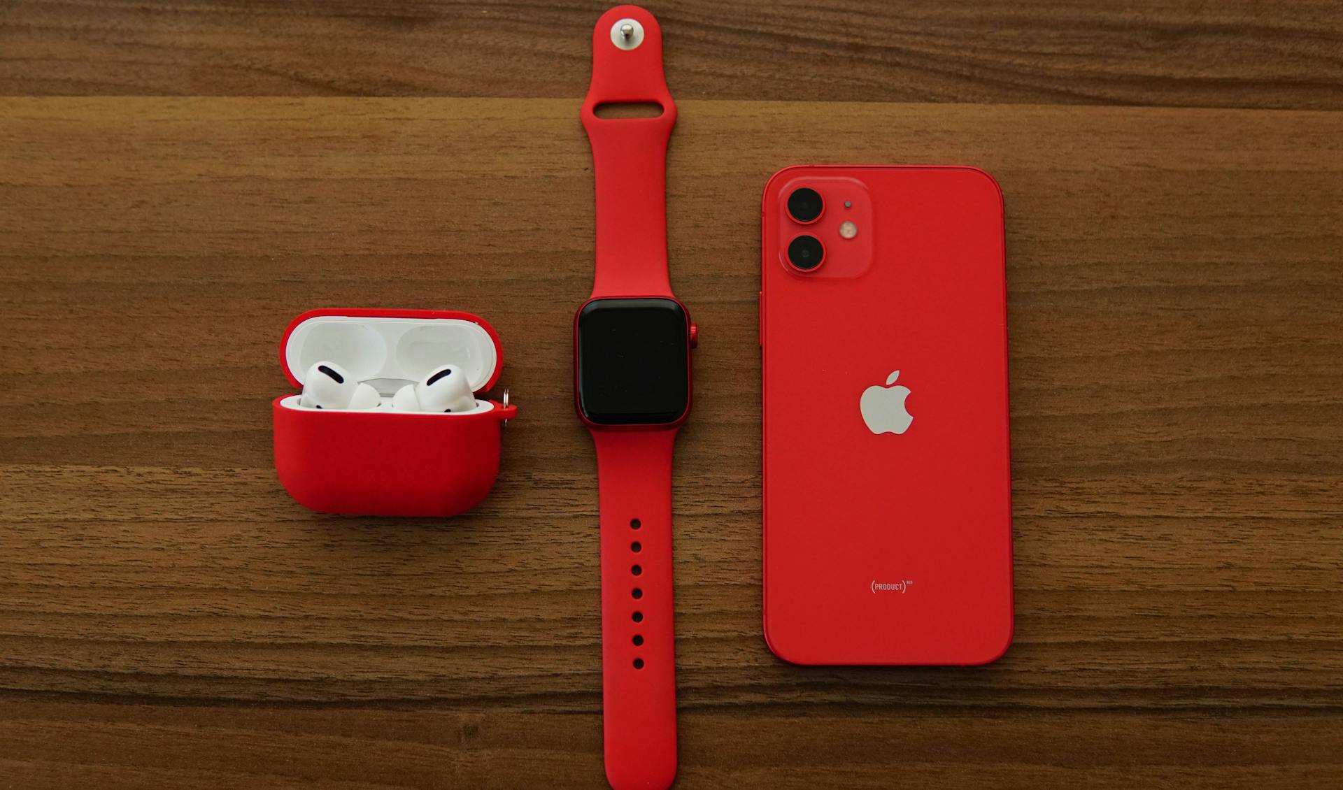 Red Apple iPhone, AirPods, and Apple Watch arranged on a wooden table.