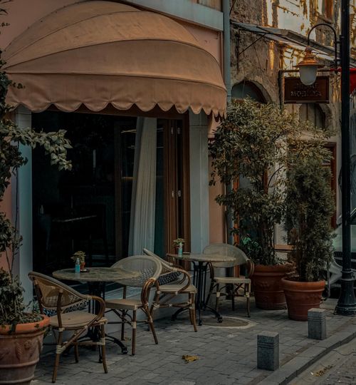 Free A picturesque outdoor café scene with tables, chairs, and potted plants on a city street. Stock Photo
