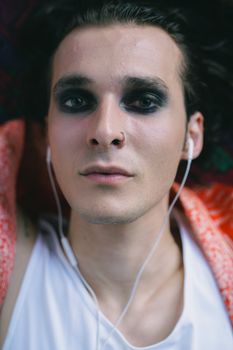 Close-up portrait of a man with dramatic eye makeup and earphones, indoors.