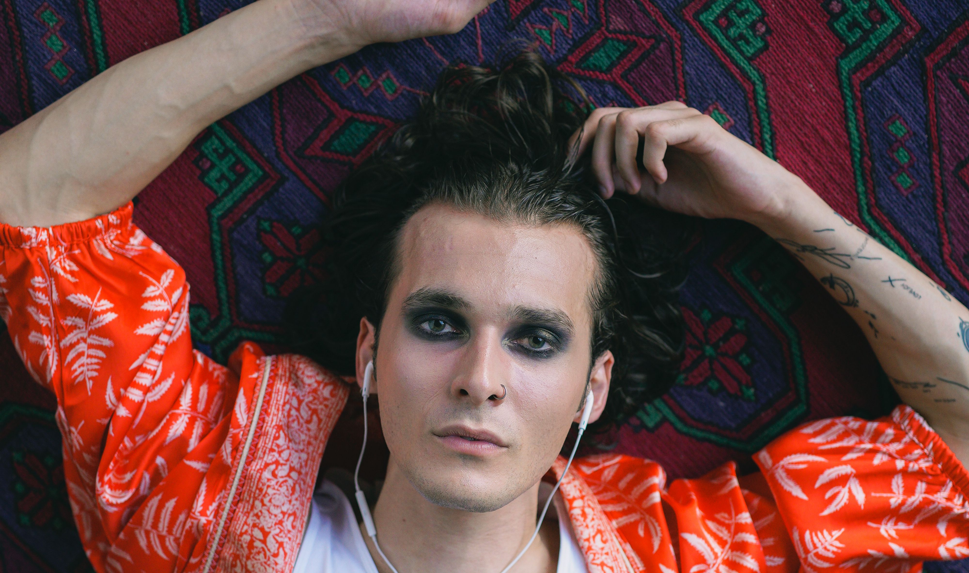 Stylish man with tattoos and make-up lying on ornate carpet, embodying modern fashion.