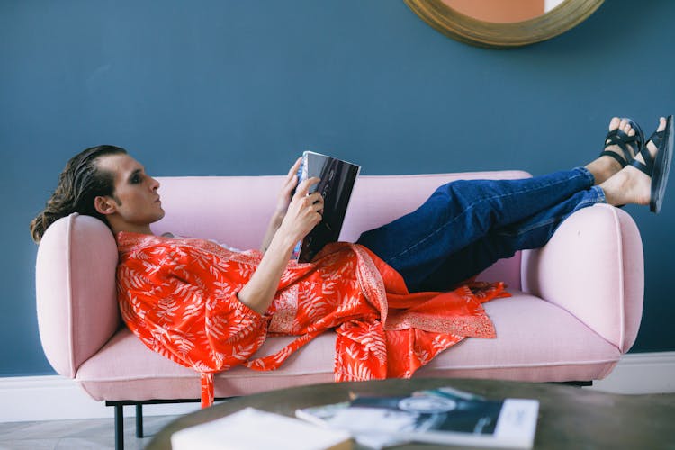 Man Lying On Back On Pink Sofa Reading Magazine