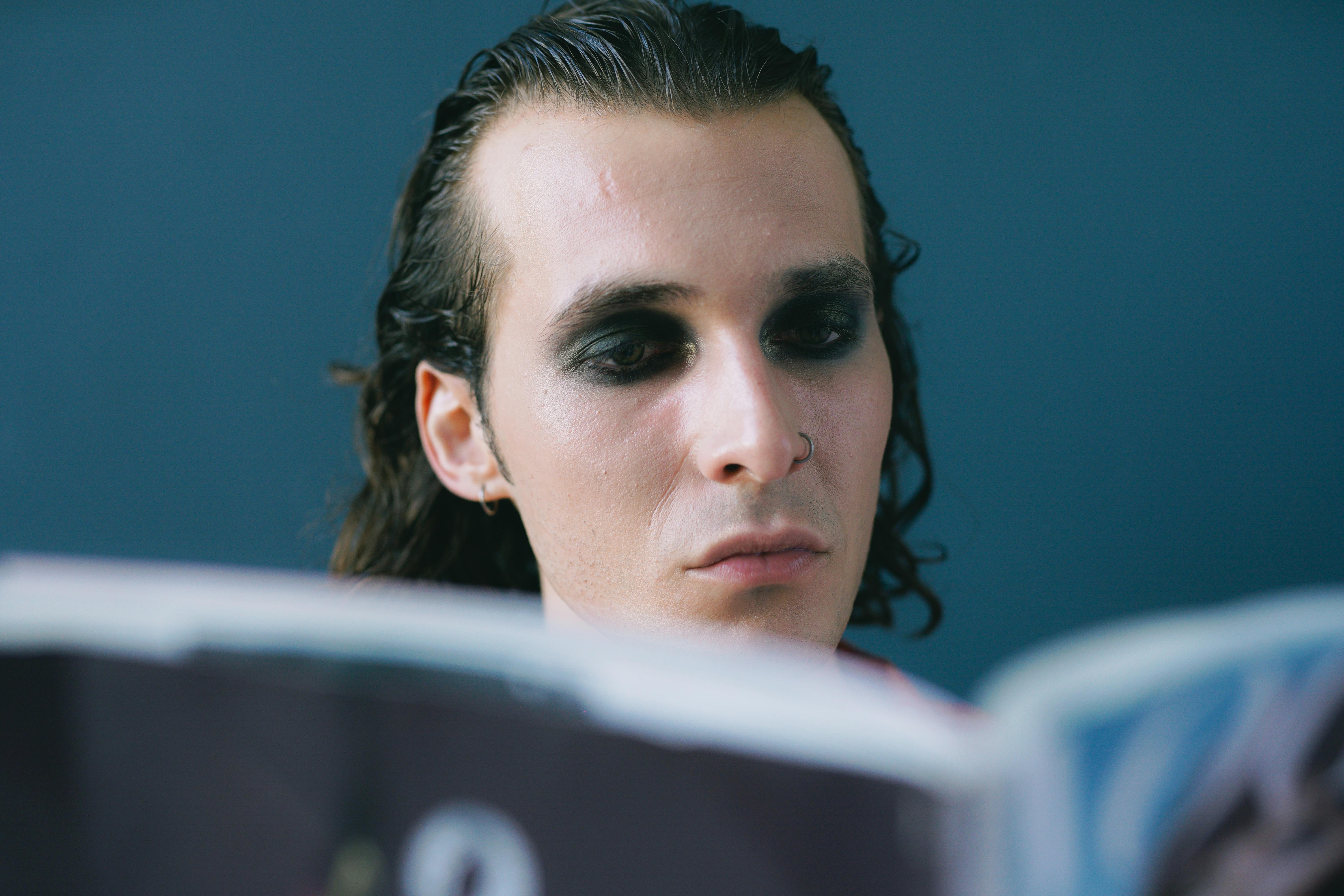 Portrait of a man with dark smokey eyeshadow, immersed in reading against a blue background.