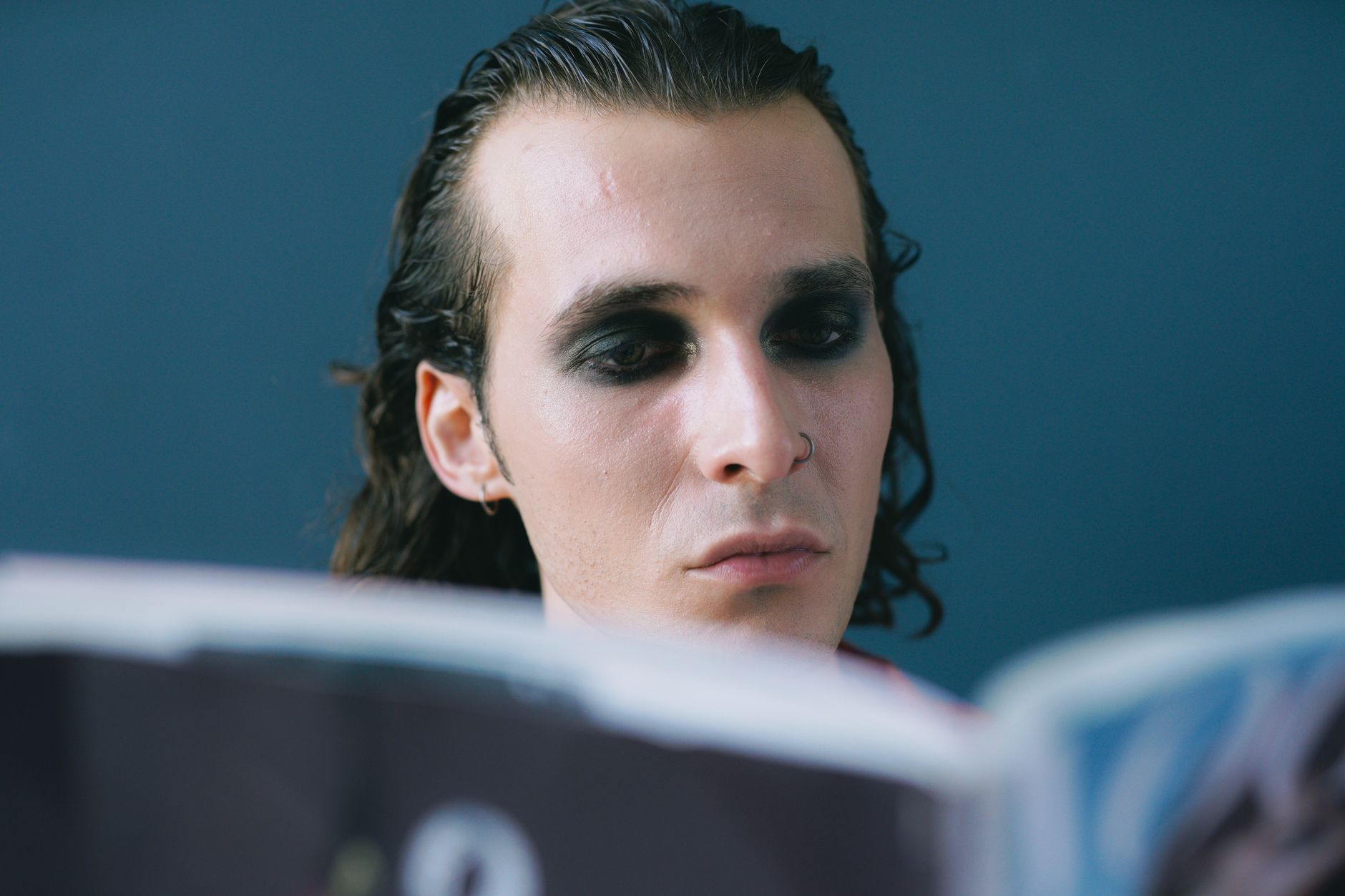 Portrait of a man with dark smokey eyeshadow, immersed in reading against a blue background.