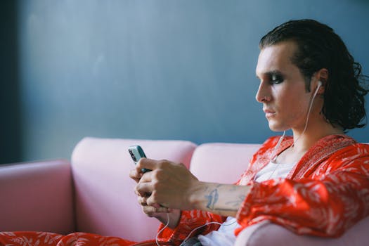A stylish man with makeup and headphones sitting on a couch indoors, using a mobile phone.