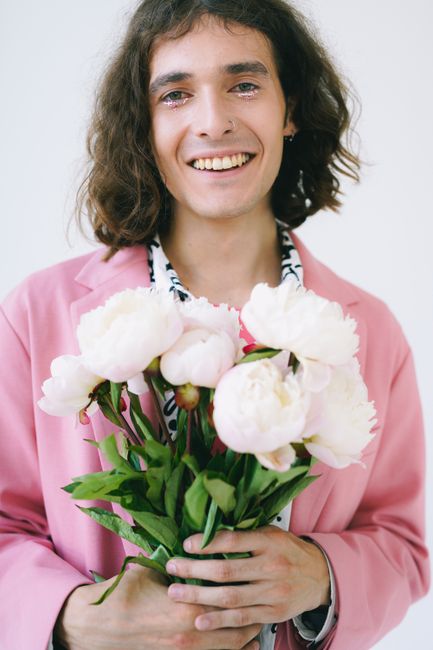 A person with a cheerful expression holds a bouquet of peonies, wearing a stylish pink jacket.