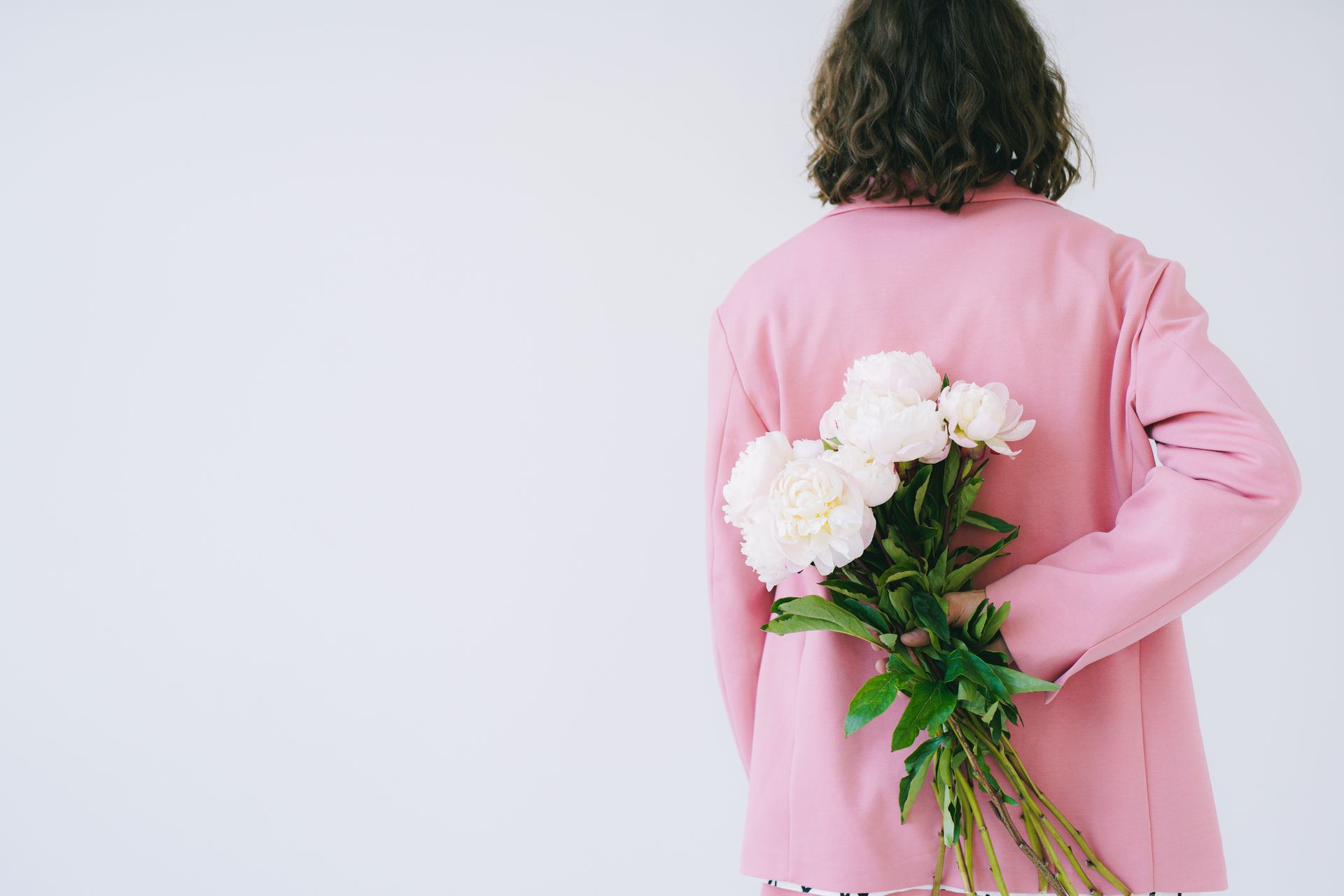 A person in a pink blazer holding a bouquet of white peonies behind their back.