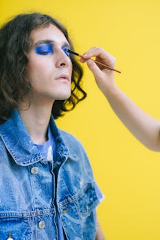 A person applies bold blue makeup to a man's eyelids against a vibrant yellow backdrop.