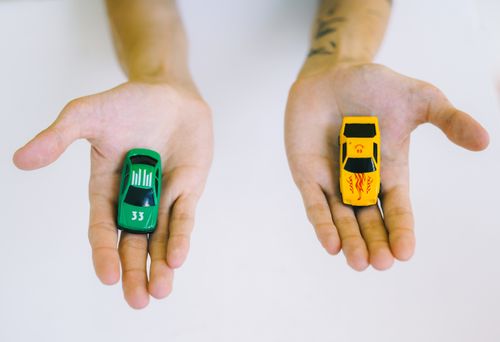 Free Pair of adult hands holding green and yellow toy cars on a white surface, symbolizing choice. Stock Photo
