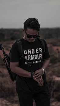 A man wearing sunglasses and backpack checks his watch in a dusk landscape.