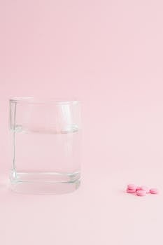 Minimalist image of a glass of water with pink pills on a pastel pink background.