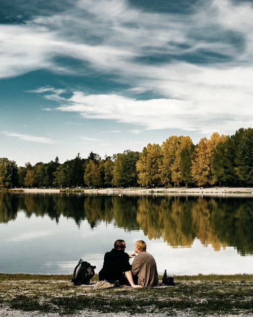 Free A couple sits by a serene lake, reflecting trees under a blue sky, enjoying nature. Stock Photo