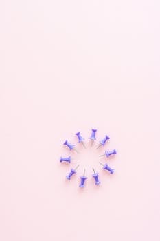 Close-up of purple push pins in a circular formation on a light pink background. Minimalist design.