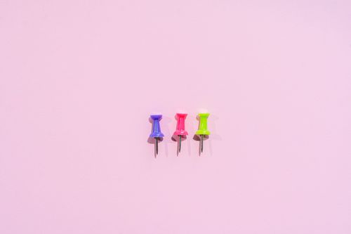 Free Three colorful push pins lined up on a pastel pink background, ideal for office themes. Stock Photo