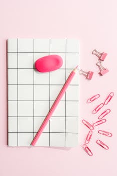 Stylish pink stationery arrangement featuring a notebook, clips, pencil, and eraser on a pink background.