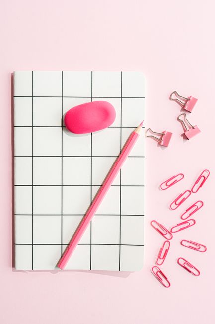 Stylish pink stationery arrangement featuring a notebook, clips, pencil, and eraser on a pink background.